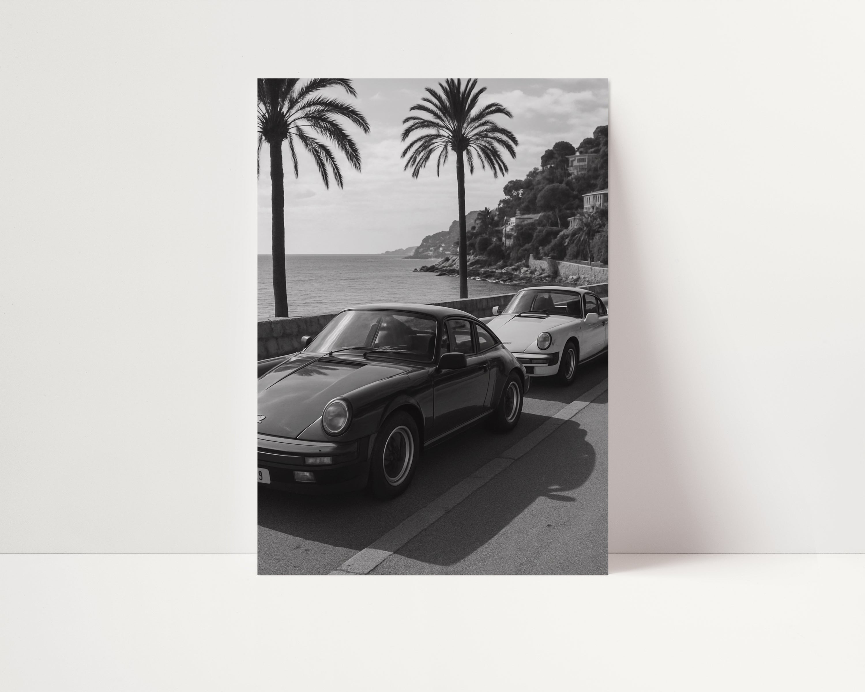 Black and white coastal photograph of two vintage sports cars parked along a palm-lined seaside road, with ocean views and hillside villas in the background, capturing timeless Riviera luxury and retro travel energy.