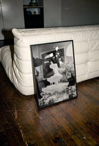 Framed black and white champagne shower poster print on wooden floor next to a white couch.