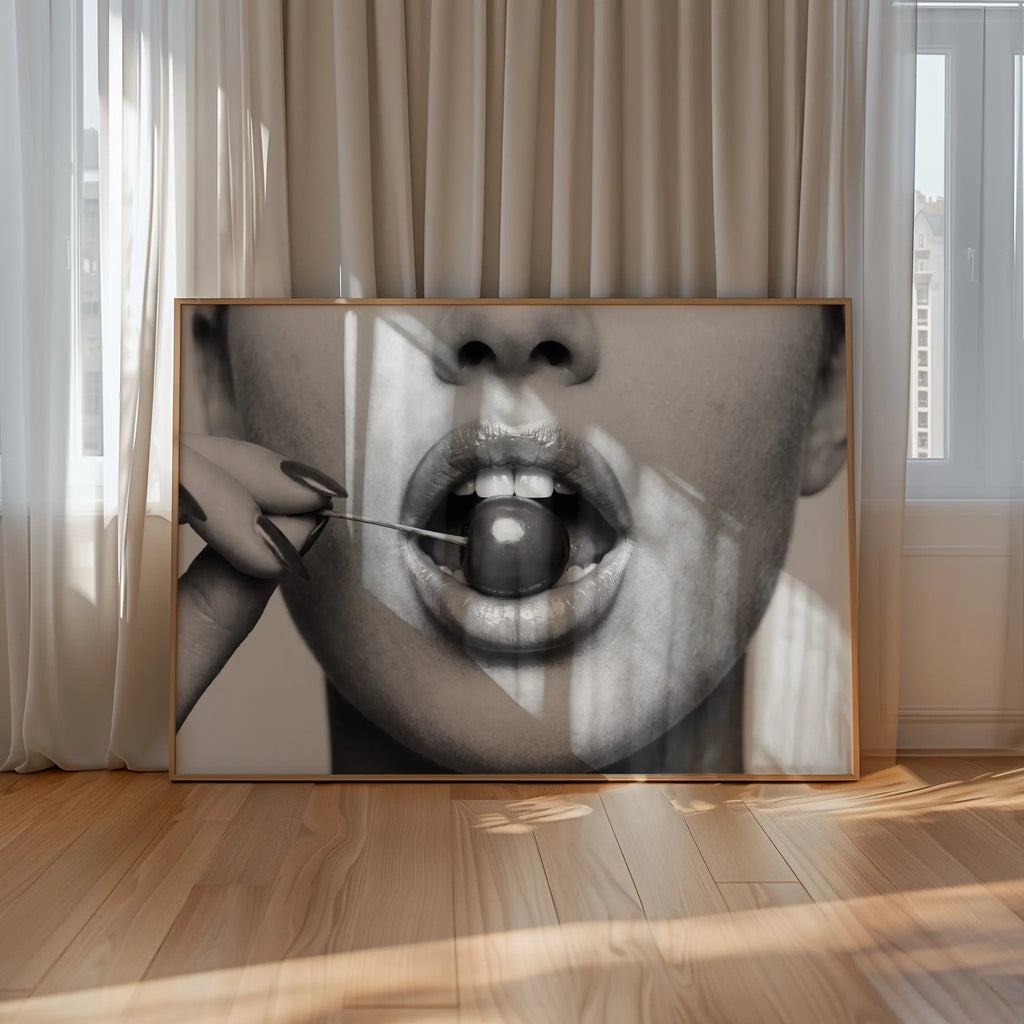 A large framed black and white art print leaning against beige curtains on a light wood floor. The artwork shows black and white poster print of cherry bomb, cherry between woman's lips framed hanging above table in dinging room with cheetah accents