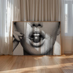 A large framed black and white art print leaning against beige curtains on a light wood floor. The artwork shows black and white poster print of cherry bomb, cherry between woman's lips framed hanging above table in dinging room with cheetah accents