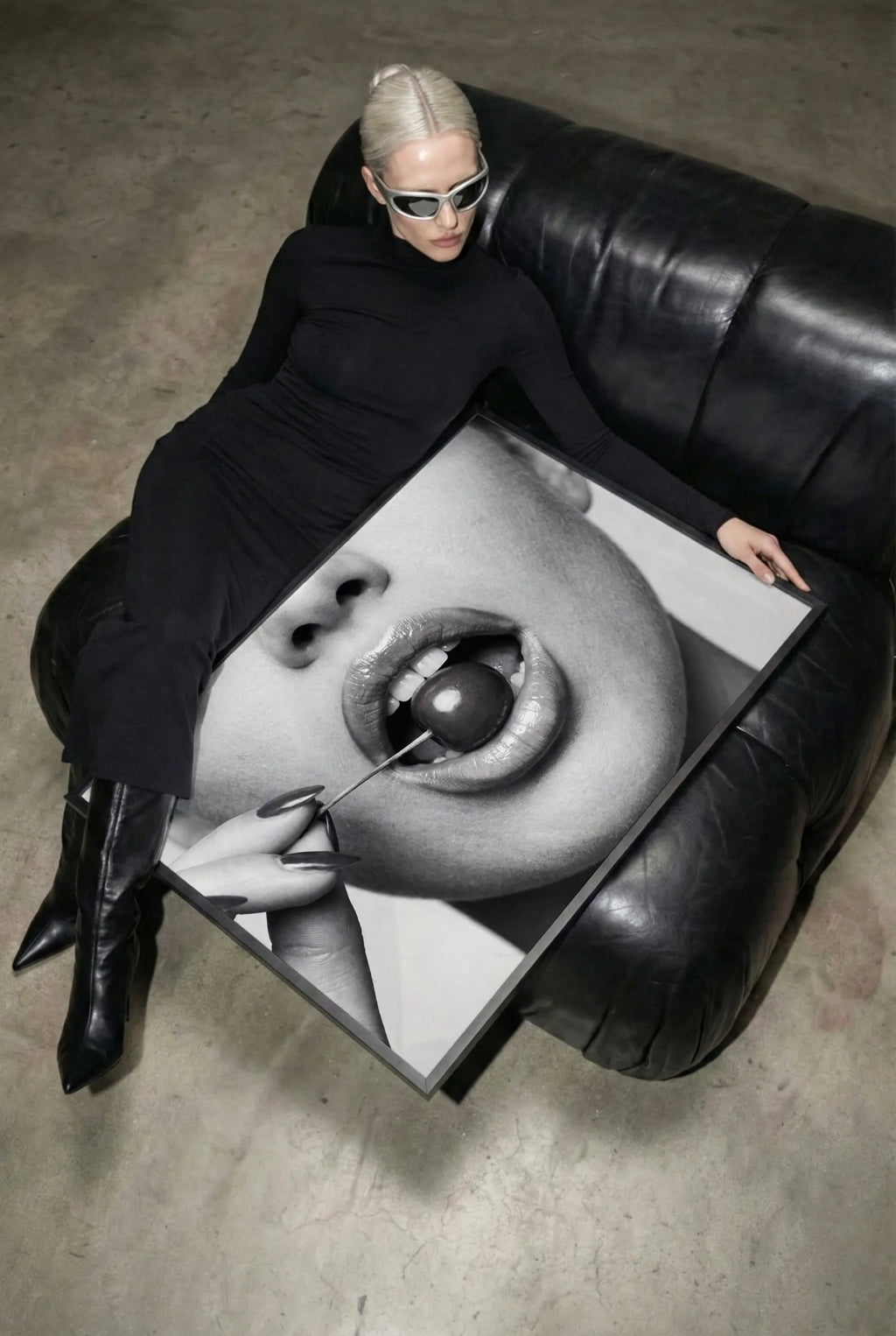 A person in a black outfit and white sunglasses reclines on a black leather sofa while holding a large black-and-white framed art print featuring a close-up of lips and a cherry.
