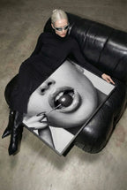 A person in a black outfit and white sunglasses reclines on a black leather sofa while holding a large black-and-white framed art print featuring a close-up of lips and a cherry.