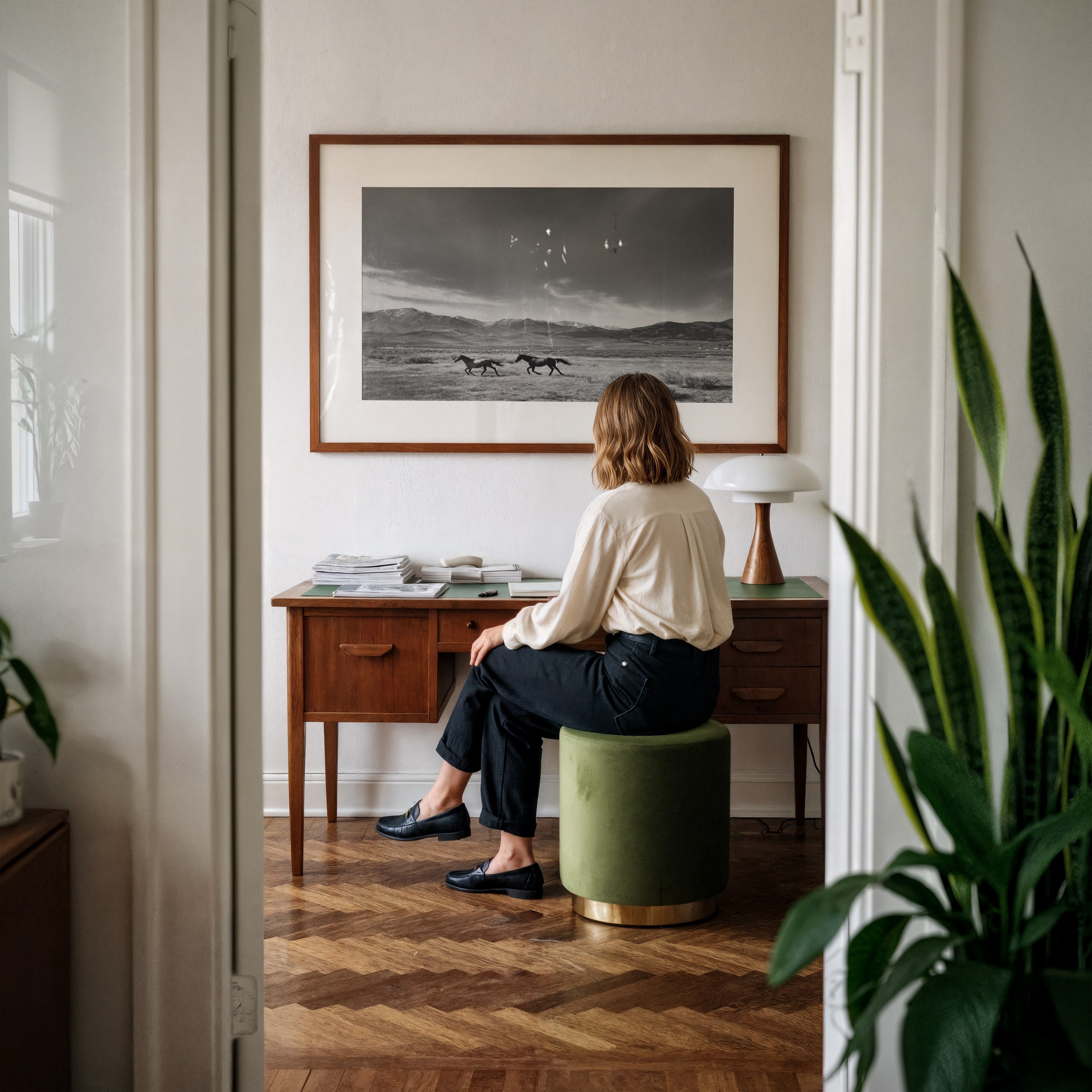 Woman sitting on a green stool in a room with a large framed black and white western wall art hanging above desk
