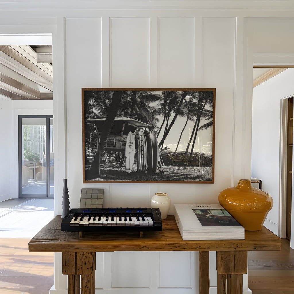 Wooden table with keyboard, books, and vase in a room with a framed black and white surfs up coastal poster print