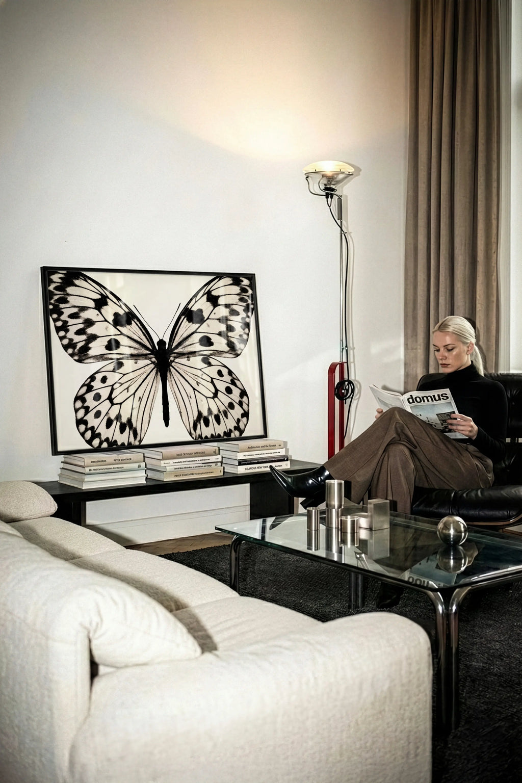 A woman sits in a black armchair reading a magazine in a modern living room, next to a glass coffee table and a large framed black-and-white butterfly artwork on a white wall.