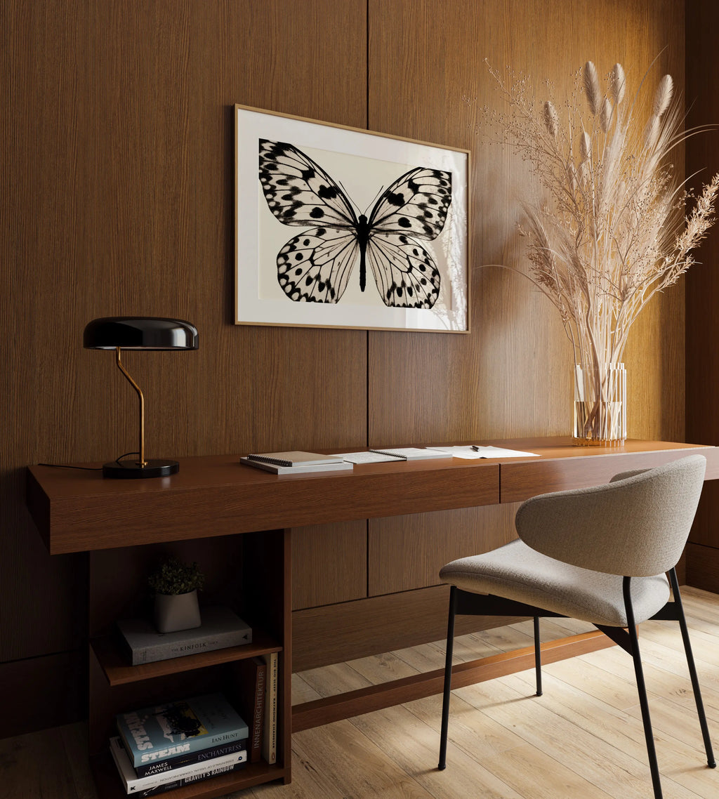 A framed black and white butterfly art print hangs on a warm wood-paneled wall above a wooden desk with a black lamp, books, and a light beige upholstered chair. A tall glass vase with dried grass stands on the right.