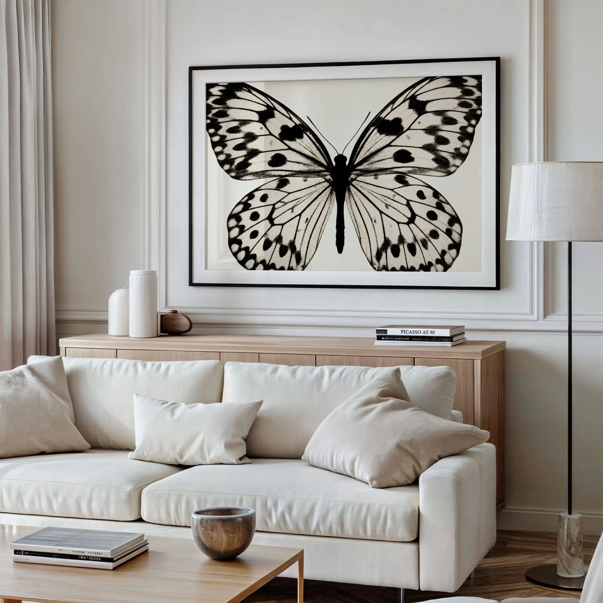 A large framed black and white butterfly art print hangs on a light beige wall above a cream sofa in a modern living room setting.