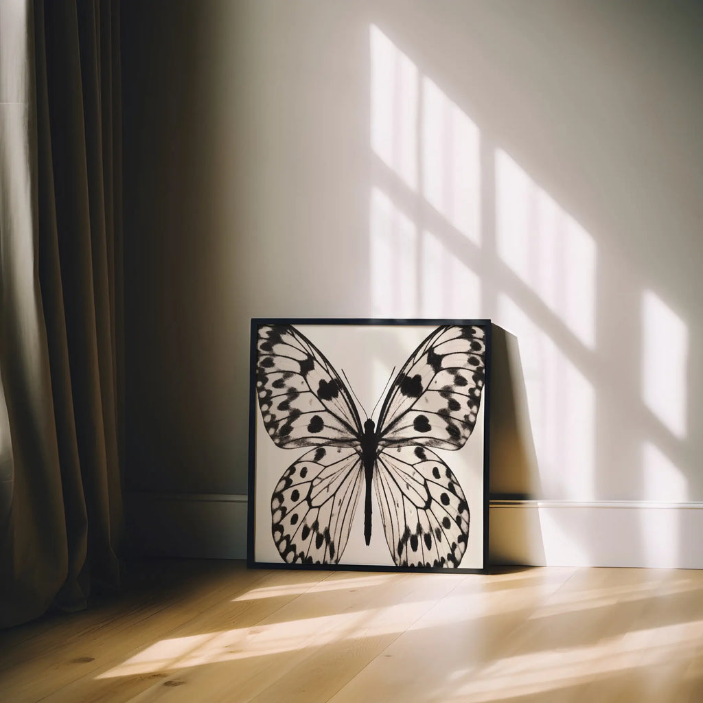 A framed butterfly art print with black and white illustration leans against a wall on a light wood floor, with warm sunlight and window shadows cast across the room.