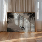 Framed artwork of a person riding a horse on a beach with palm trees, displayed in a room with curtains and wooden floor.