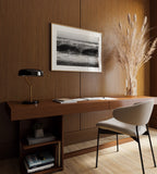 Modern home office with wooden desk, chair, lamp, and framed black and white high tide poster print on a wooden wall.