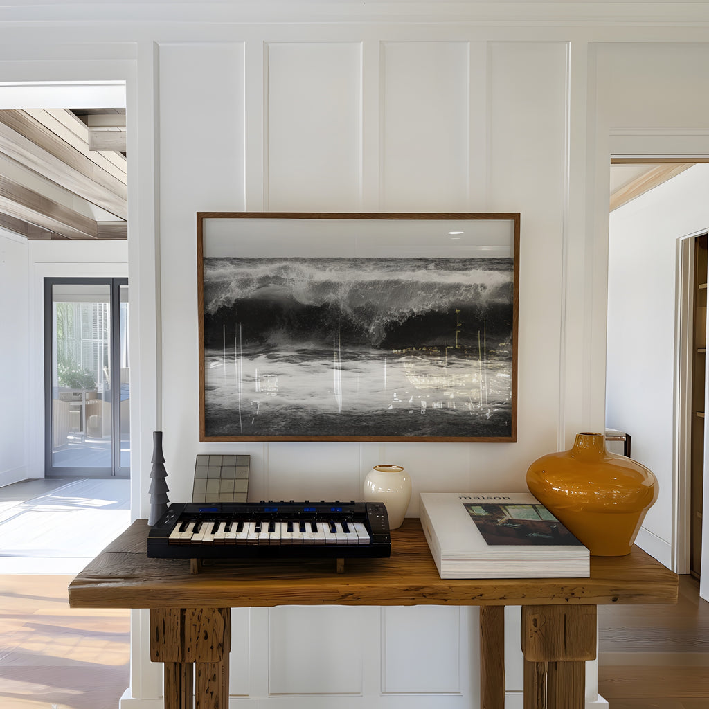 Music keyboard on a wooden table with a framed black and white high tide poster print above, in a bright room.
