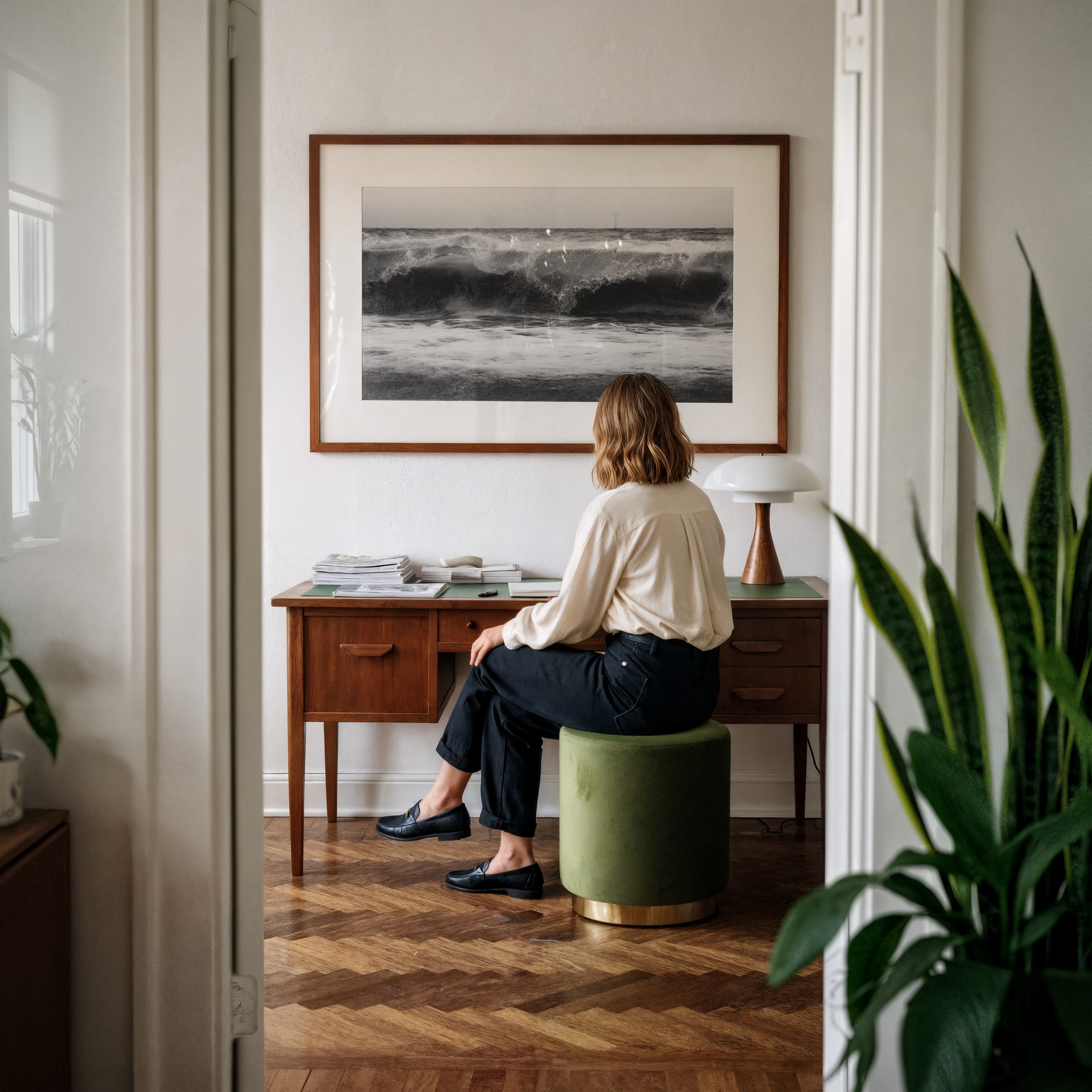 Person sitting on a green stool in front of a wooden desk with aframed black and white high tide poster print  on the wall.