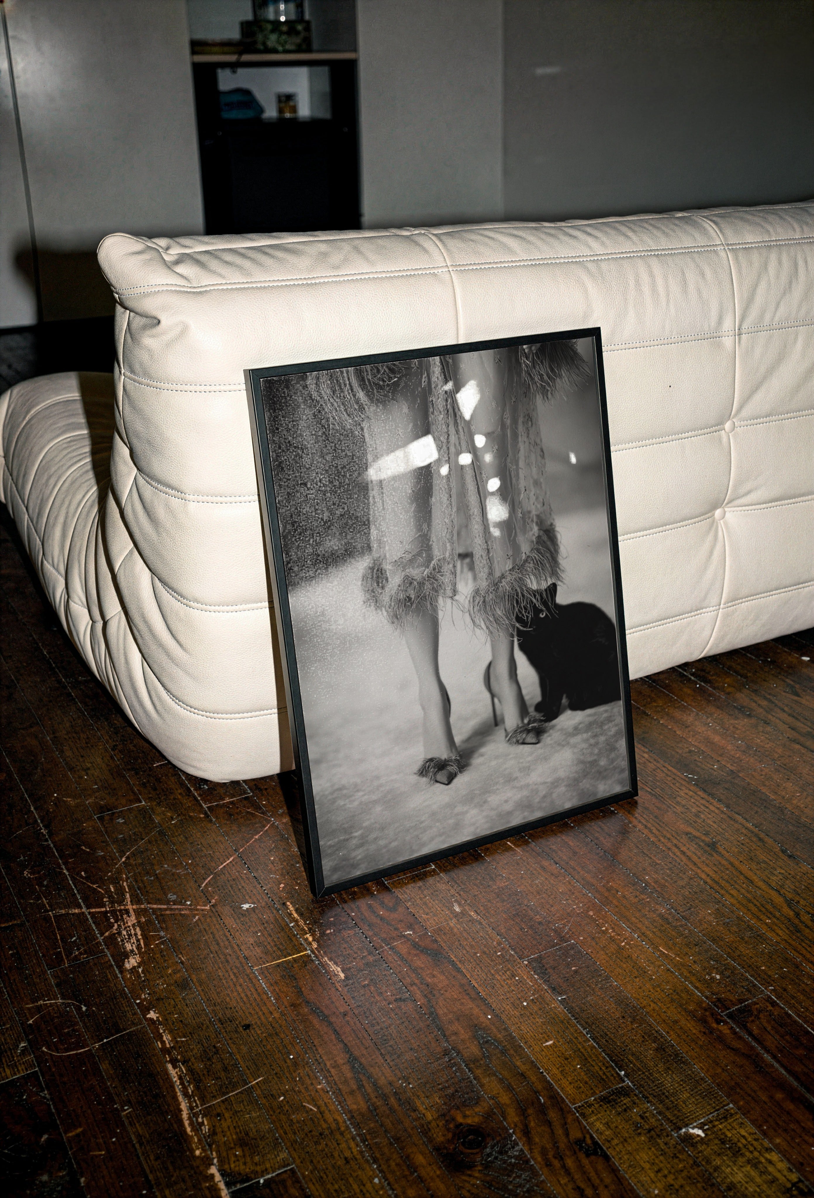 framed black and white kitty kitty poster print on a wooden floor next to a white couch.