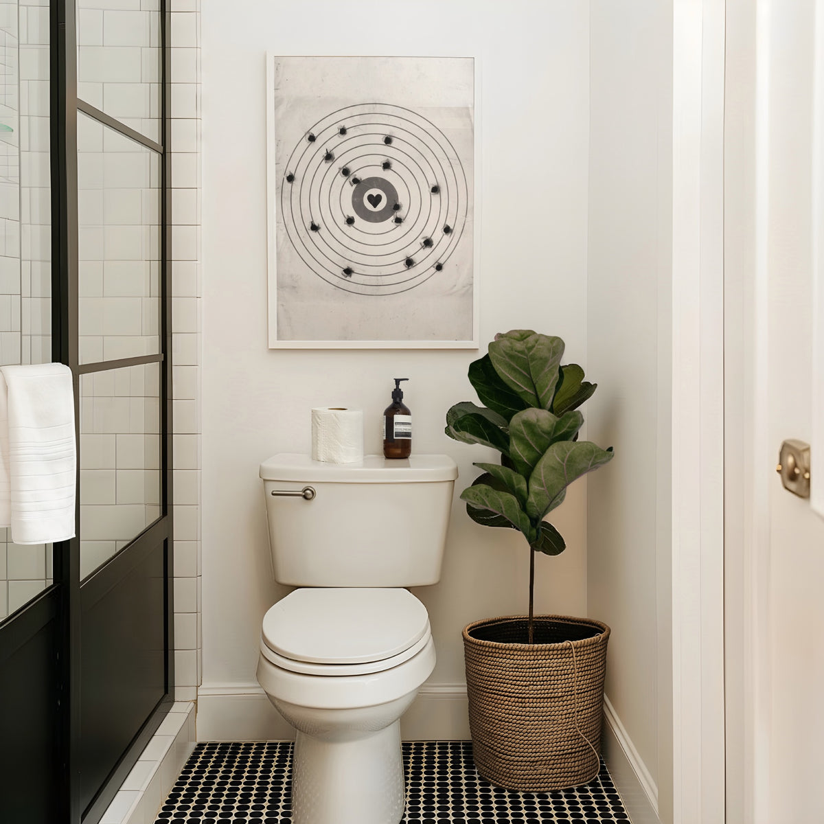 A framed moving target wall art print featuring a black bullseye with a heart at the center hanging above toilet in bathroom