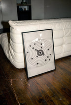 A framed moving target wall art print featuring a black bullseye with a heart at the center, leaning against a white tufted sofa on a dark wood floor.
