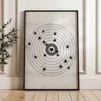 A framed moving target wall art print featuring a black bullseye with a heart at the center, leaning against a white paneled wall on a light wood floor next to a green plant.