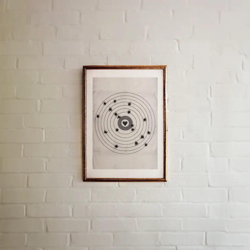 A framed moving target wall art print featuring a black bullseye with a heart at the center, hanging on a white painted brick wall.