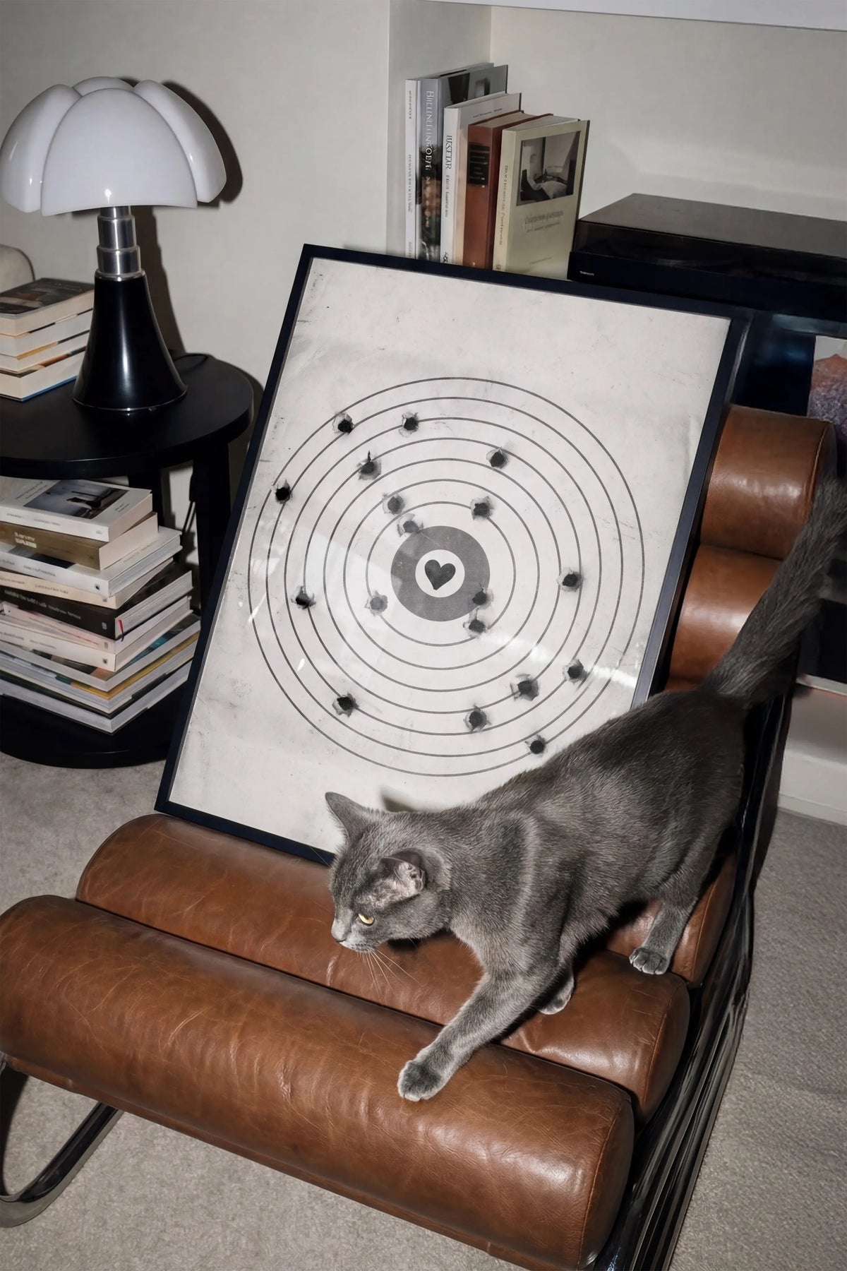 A gray cat walks across a brown leather armchair next to a framed moving target wall art print featuring a black bullseye with a heart at the center