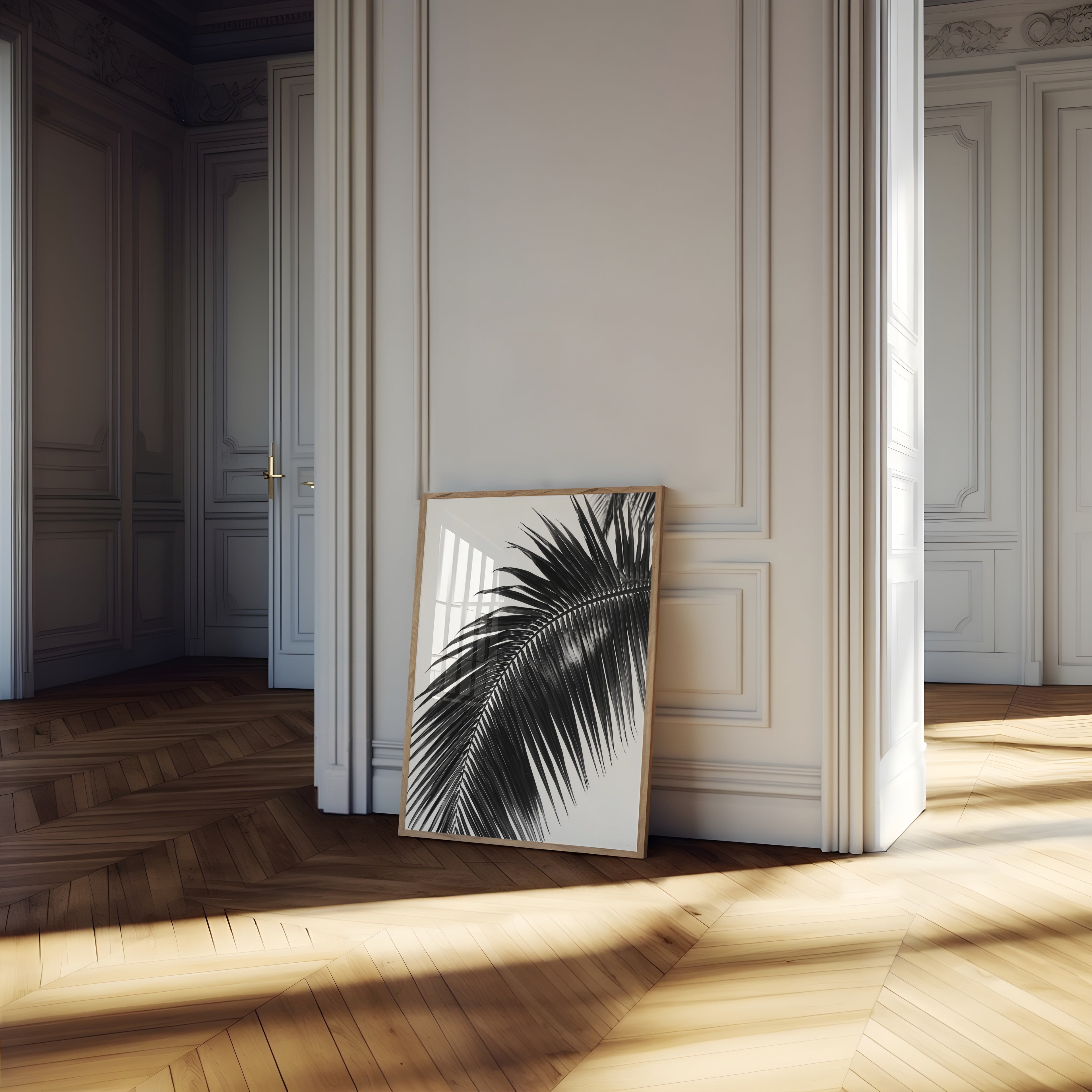 A framed black and white palm leaf art print leans against a cream paneled wall on a light wood herringbone floor in a sunlit room.