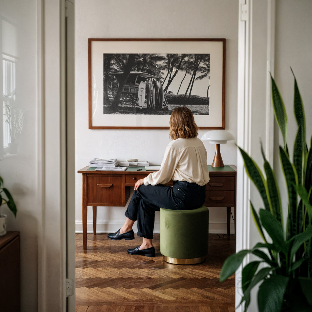 Person sitting on a green stool in a room with a wooden desk and Framed black and white Surfs Up poster print 